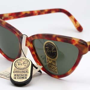 May include: Tortoiseshell Ray-Ban sunglasses with green lenses. The sunglasses have a gold Ray-Ban logo on the right temple and a gold tag with the text "Ray-Ban 5-15' Lens XLT" on the right lens. The sunglasses also have a black tag with the text "Original Bausch & Lomb" attached to the left temple.