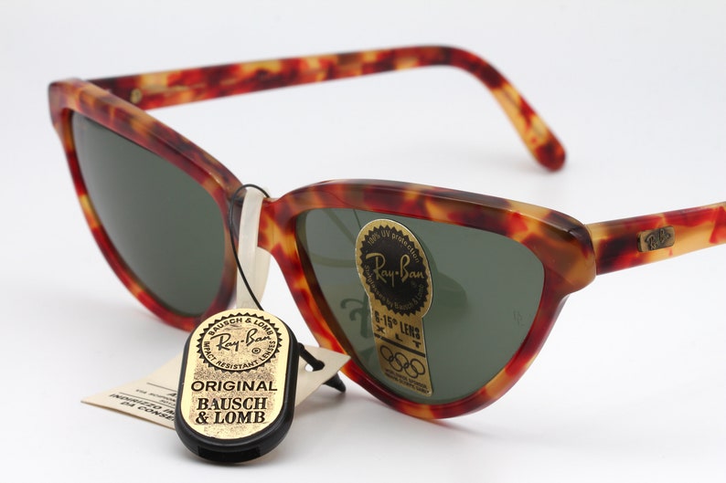 May include: Tortoiseshell Ray-Ban sunglasses with green lenses. The sunglasses have a gold Ray-Ban logo on the right temple and a gold tag with the text "Ray-Ban 5-15' Lens XLT" on the right lens. The sunglasses also have a black tag with the text "Original Bausch & Lomb" attached to the left temple.