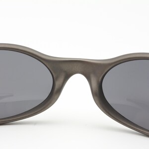 May include: Close-up of a pair of oval-shaped sunglasses with dark gray lenses and a matte gray frame. The sunglasses have a sleek, modern design, suitable for outdoor activities.