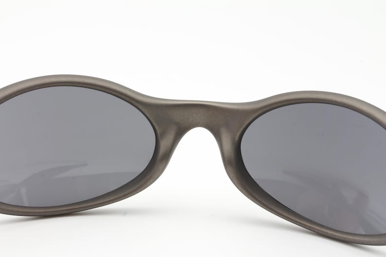 May include: Close-up of a pair of oval-shaped sunglasses with dark gray lenses and a matte gray frame. The sunglasses have a sleek, modern design, suitable for outdoor activities.