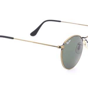 Puede incluir: Gafas de sol redondas doradas con lentes verdes y patillas negras. Las gafas tienen el logotipo de Ray-Ban en la lente derecha.