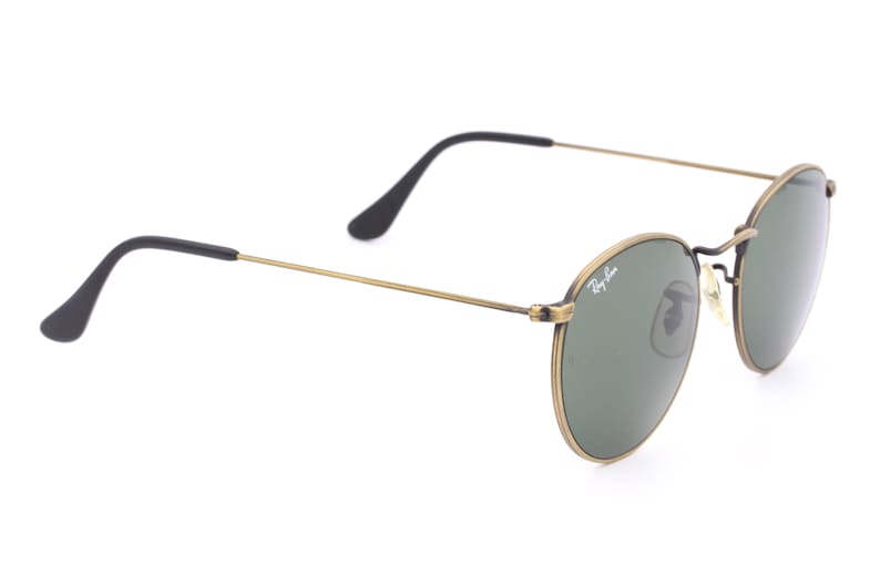 Puede incluir: Gafas de sol redondas doradas con lentes verdes y patillas negras. Las gafas tienen el logotipo de Ray-Ban en la lente derecha.