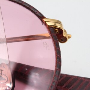 May include: Close-up of a pair of pink sunglasses with a gold metal frame. The sunglasses are folded and the frame is visible. The sunglasses are in a burgundy leather case.