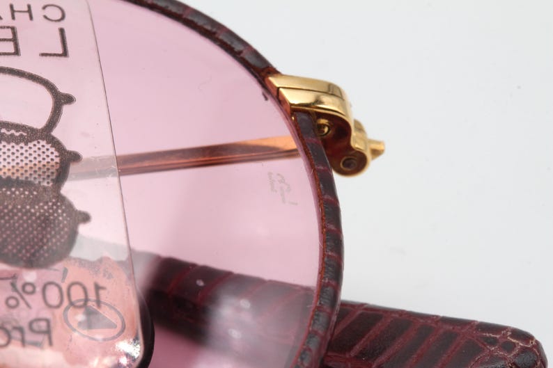 May include: Close-up of a pair of pink sunglasses with a gold metal frame. The sunglasses are folded and the frame is visible. The sunglasses are in a burgundy leather case.