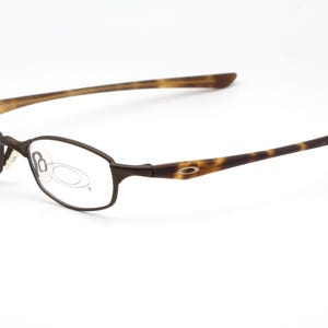 May include: Brown metal-framed eyeglasses with oval lenses. The temples have a tortoise shell pattern. The glasses are set against a white background. The Oakley logo is visible on the temple.