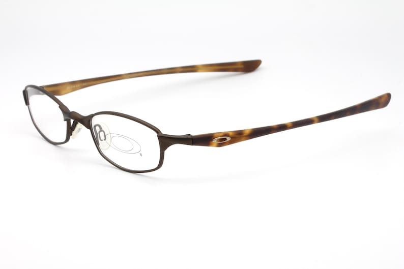 May include: Brown metal-framed eyeglasses with oval lenses. The temples have a tortoise shell pattern. The glasses are set against a white background. The Oakley logo is visible on the temple.