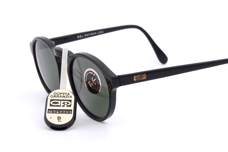 May include: Black round Ray-Ban sunglasses with green lenses. The sunglasses have a gold Ray-Ban logo on the right arm and the text "B&L Ray-Ban USA" on the left arm.