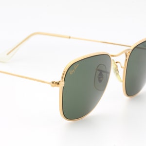 May include: Gold-framed sunglasses with green lenses. The sunglasses have a classic, square shape and are made by Ray-Ban.