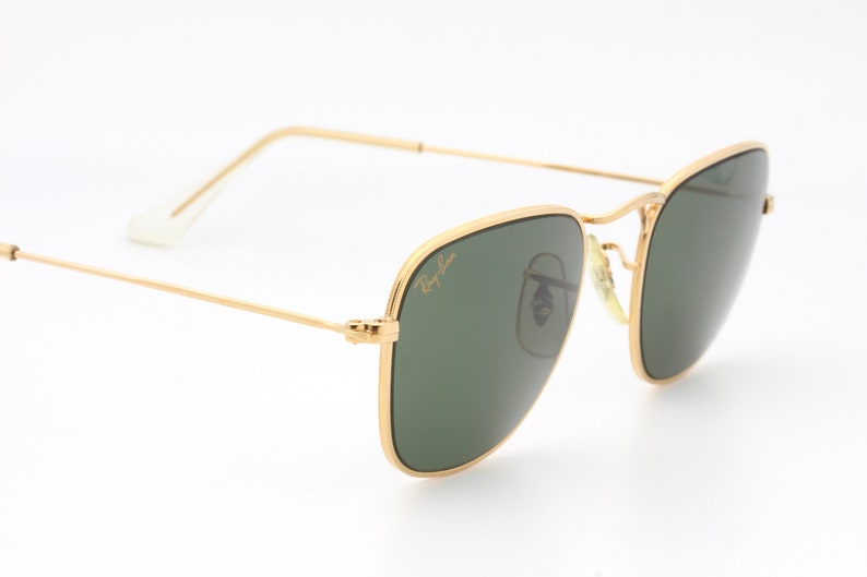 May include: Gold-framed sunglasses with green lenses. The sunglasses have a classic, square shape and are made by Ray-Ban.