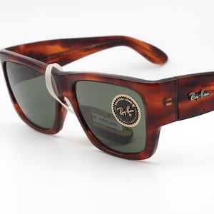 Puede incluir: Gafas de sol carey marr&oacute;n con lentes oscuras. El logotipo de Ray-Ban es visible en la patilla. Una banda blanca asegura las gafas. Las lentes tienen un tinte verde. Gafas de sol nuevas con etiqueta de precio.