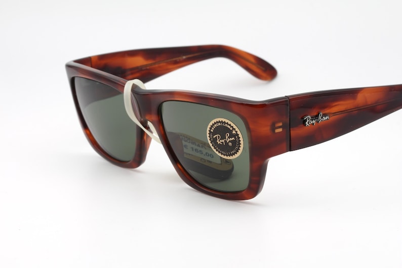 Puede incluir: Gafas de sol carey marr&oacute;n con lentes oscuras. El logotipo de Ray-Ban es visible en la patilla. Una banda blanca asegura las gafas. Las lentes tienen un tinte verde. Gafas de sol nuevas con etiqueta de precio.