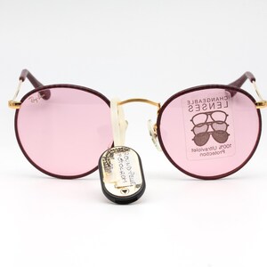 May include: Round burgundy sunglasses with pink lenses. The sunglasses have gold accents and the text "CHANGEABLE LENSES" is printed on the right lens.