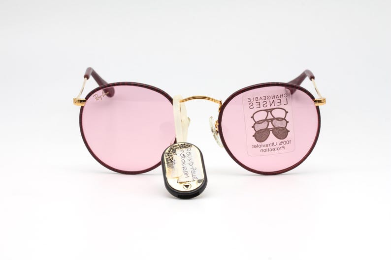 May include: Round burgundy sunglasses with pink lenses. The sunglasses have gold accents and the text "CHANGEABLE LENSES" is printed on the right lens.