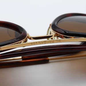 May include: Close-up of sunglasses with brown lenses and a tortoiseshell frame. The gold-colored metal frame has the text "B&L RAY-BAN USA" etched on the side. The arms of the sunglasses are a dark brown color.