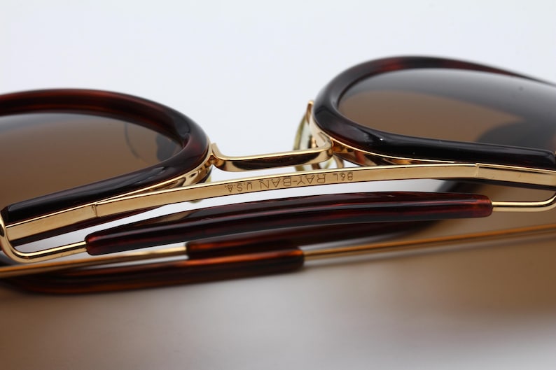 May include: Close-up of sunglasses with brown lenses and a tortoiseshell frame. The gold-colored metal frame has the text "B&L RAY-BAN USA" etched on the side. The arms of the sunglasses are a dark brown color.