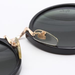 May include: Black round sunglasses with gold accents and a clear plastic nose bridge. The sunglasses have a dark green lens.