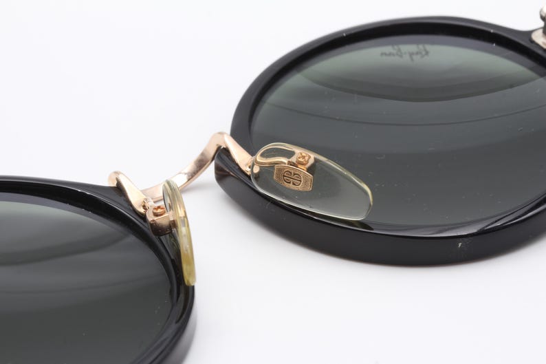 May include: Black round sunglasses with gold accents and a clear plastic nose bridge. The sunglasses have a dark green lens.