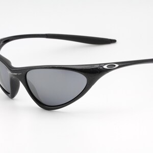 Puede incluir: Gafas de sol negras con lentes grises. Las gafas tienen una montura y patillas curvas, con un peque&ntilde;o logotipo plateado en las patillas. Las lentes son de color gris oscuro y la montura es negra texturizada.