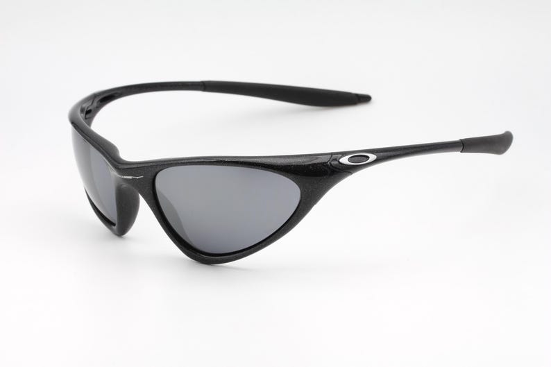 Puede incluir: Gafas de sol negras con lentes grises. Las gafas tienen una montura y patillas curvas, con un peque&ntilde;o logotipo plateado en las patillas. Las lentes son de color gris oscuro y la montura es negra texturizada.