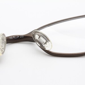 May include: Close-up of brown eyeglasses with clear lenses. The glasses have a thin, curved frame and clear nose pads. The frame is marked with the numbers 49 and 20. The glasses are set against a white background.