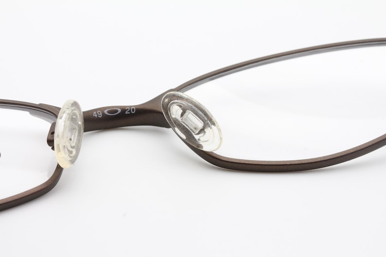 May include: Close-up of brown eyeglasses with clear lenses. The glasses have a thin, curved frame and clear nose pads. The frame is marked with the numbers 49 and 20. The glasses are set against a white background.