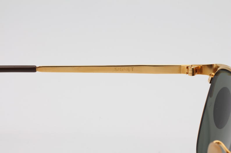 May include: Gold-tone metal sunglasses with green lenses. The sunglasses have a Signet inscription on the arm.
