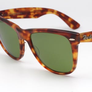 May include: Tortoiseshell-patterned sunglasses with green lenses. The frames are a mix of brown and amber hues. The Ray-Ban logo is visible on the right temple. A classic eyewear accessory.