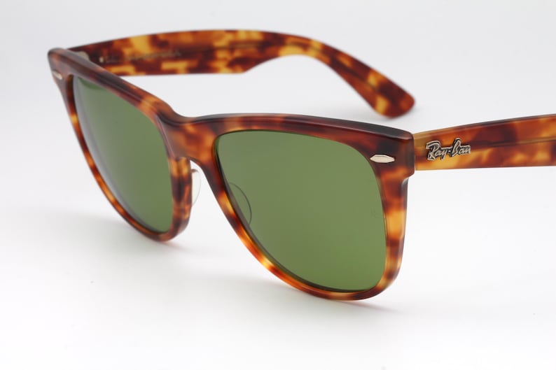 May include: Tortoiseshell-patterned sunglasses with green lenses. The frames are a mix of brown and amber hues. The Ray-Ban logo is visible on the right temple. A classic eyewear accessory.