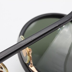 May include: Black sunglasses with green lenses and a gold metal frame. The sunglasses have a thick black plastic frame and a thin gold metal band around the lenses.