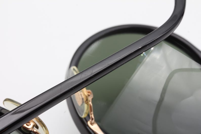 May include: Black sunglasses with green lenses and a gold metal frame. The sunglasses have a thick black plastic frame and a thin gold metal band around the lenses.