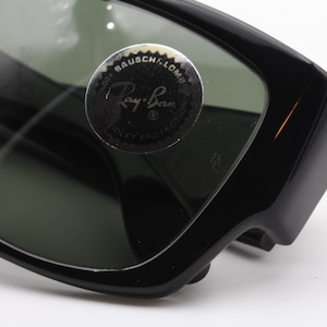 May include: Close-up of black Ray-Ban sunglasses. The sunglasses have a thick, rectangular frame and dark green lenses. A circular sticker with the Ray-Ban logo is visible on the lens. The sunglasses are a classic style.