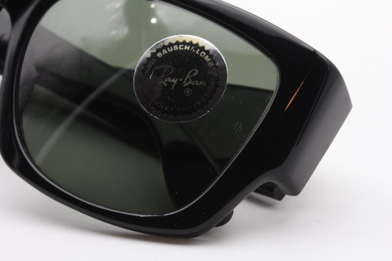 May include: Close-up of black Ray-Ban sunglasses. The sunglasses have a thick, rectangular frame and dark green lenses. A circular sticker with the Ray-Ban logo is visible on the lens. The sunglasses are a classic style.