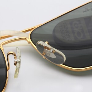 May include: Gold-tone aviator sunglasses with dark gray lenses. The sunglasses have a white plastic nose bridge and a gold-tone metal frame. The right lens has a small, round sticker with the text "COPPIA GARANZIA" in black.