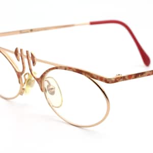 May include: Gold-tone metal eyeglasses with a red accent on the temple arm. The frame has a mottled brown and gold finish. The glasses have a round lens shape.