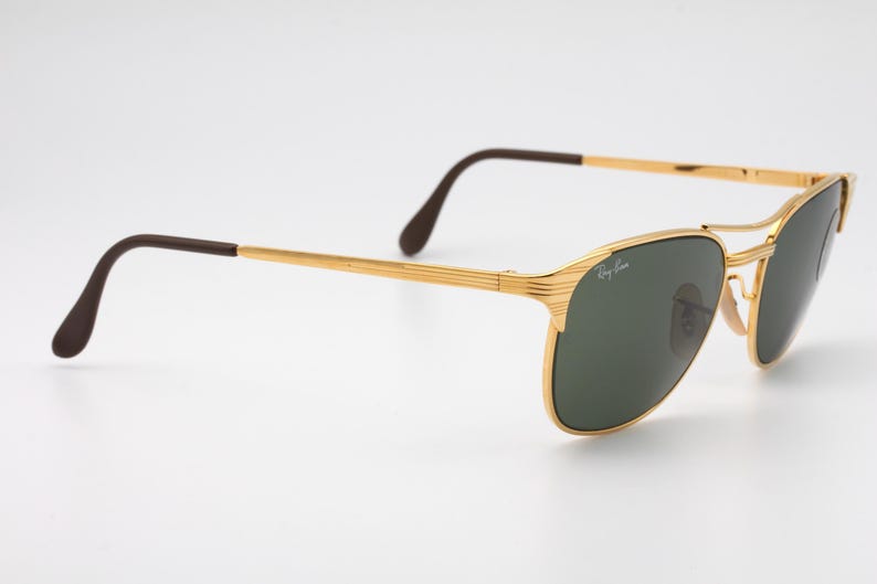 May include: Gold-tone aviator sunglasses with green lenses and brown temple tips. The sunglasses have a Ray-Ban logo on the right lens.