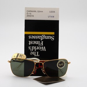 May include: Gold-framed sunglasses with green lenses. The sunglasses are sitting on a white surface with a black and white card that reads "The World's Finest Sunglasses".