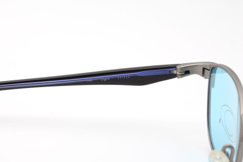 May include: Close-up of a pair of eyeglasses with a silver frame and blue lenses. The temples are black with the text "140 Light 11-717". The lenses are a light blue color.