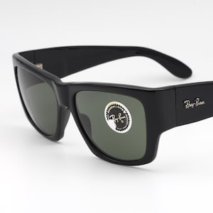 May include: Black Ray-Ban sunglasses with a square frame and dark lenses. The sunglasses have the Ray-Ban logo on the temple and a sticker on the lens. The sunglasses are on a white background.