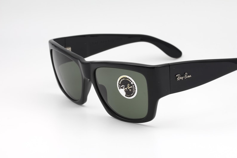 May include: Black Ray-Ban sunglasses with a square frame and dark lenses. The sunglasses have the Ray-Ban logo on the temple and a sticker on the lens. The sunglasses are on a white background.