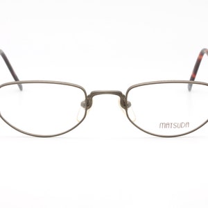 Può includere: Un paio di occhiali in metallo color argento con montatura rettangolare e lenti trasparenti. Il nome del marchio "MATSUDA" è stampato sulla lente destra.
