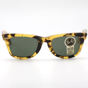 Puede incluir: Gafas de sol Ray-Ban cl&aacute;sicas con montura con estampado de carey y lentes verde oscuro. Las gafas tienen forma rectangular y una peque&ntilde;a pegatina con el logotipo de Ray-Ban y los anillos ol&iacute;mpicos.