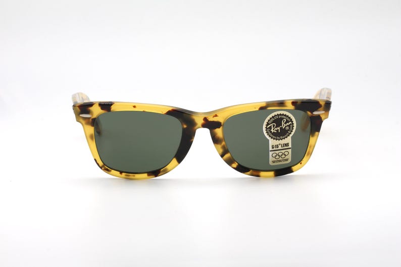 Puede incluir: Gafas de sol Ray-Ban cl&aacute;sicas con montura con estampado de carey y lentes verde oscuro. Las gafas tienen forma rectangular y una peque&ntilde;a pegatina con el logotipo de Ray-Ban y los anillos ol&iacute;mpicos.