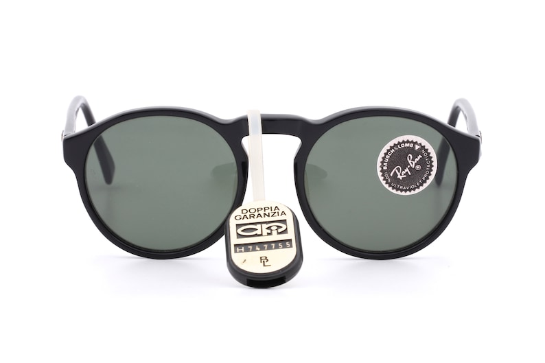 May include: Black round sunglasses with green lenses. The sunglasses have a Ray-Ban logo on the right lens and a tag that says "Doppia Garanzia" with a serial number.