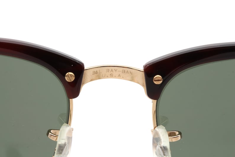 Może przedstawiać: Zbliżenie na złoty metalowy mostek brązowych okular&oacute;w przeciwsłonecznych Ray-Ban w kolorze ż&oacute;łwim. Mostek jest grawerowany napisem "B&L RAY-BAN U.S.A."