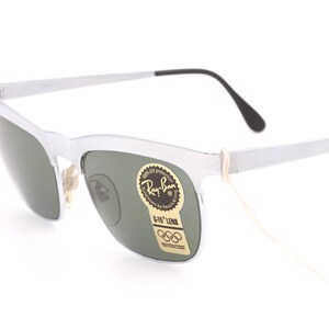 May include: Silver Ray-Ban sunglasses with green lenses and a G-15 lens label. The sunglasses have a thin, white string attached to the frame.