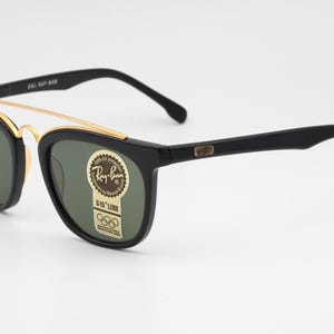 May include: Black Ray-Ban sunglasses with a gold-colored brow bar and green-tinted lenses. The frames have a classic design with a rectangular shape. The sunglasses have a sticker with the Ray-Ban logo and Olympic rings.