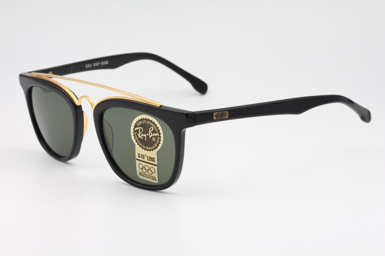 May include: Black Ray-Ban sunglasses with a gold-colored brow bar and green-tinted lenses. The frames have a classic design with a rectangular shape. The sunglasses have a sticker with the Ray-Ban logo and Olympic rings.