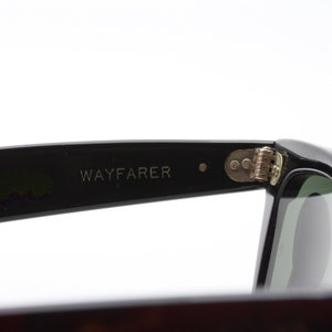 May include: Close-up of the black Wayfarer sunglasses with a silver hinge. The sunglasses have a green lens.