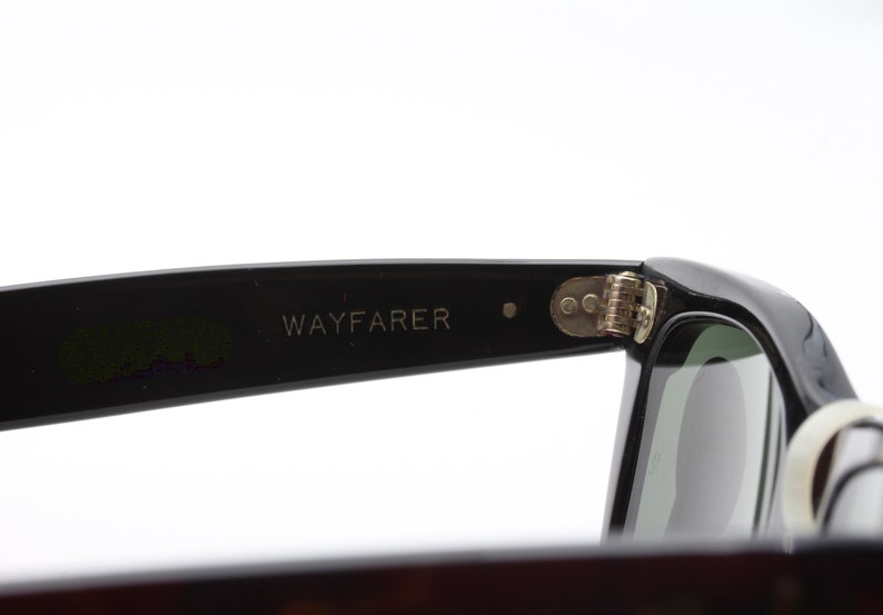 May include: Close-up of the black Wayfarer sunglasses with a silver hinge. The sunglasses have a green lens.