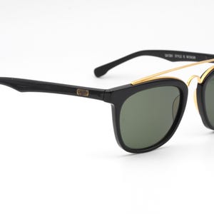 May include: Black sunglasses with a gold-colored metal bridge and dark green lenses. The sunglasses have a classic design with a rectangular frame and a curved temple. The text "Gatsby Style & Woman" is visible on the frame.